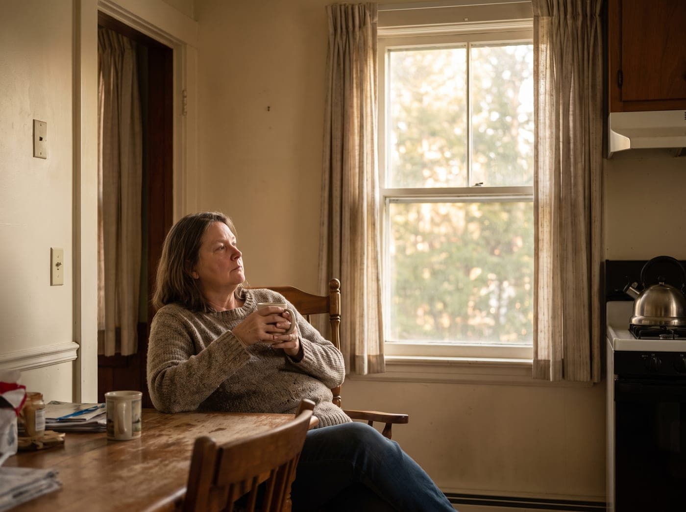 A woman sitting at a kitchen table with a coffee in early morning light